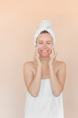 Young woman with problem skin applies wash foam to her face. Girl with acne in towel on a beige background.