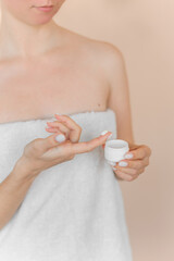 Woman in a bath towel applies cream from a bottle to her finger. Close up without face on a beige background.