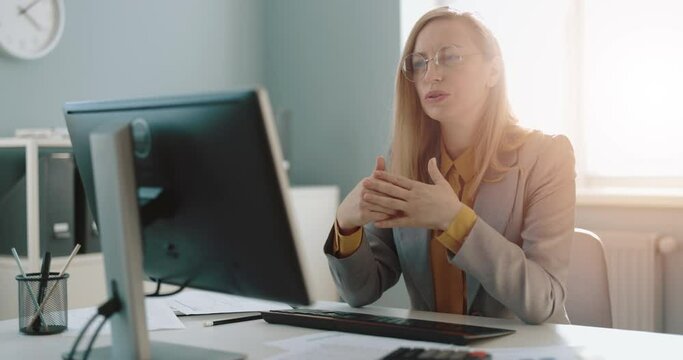 Mature woman in formal wear sitting at office desk, talking and gesturing during conference call on modern pc. Business conversation and online meeting.