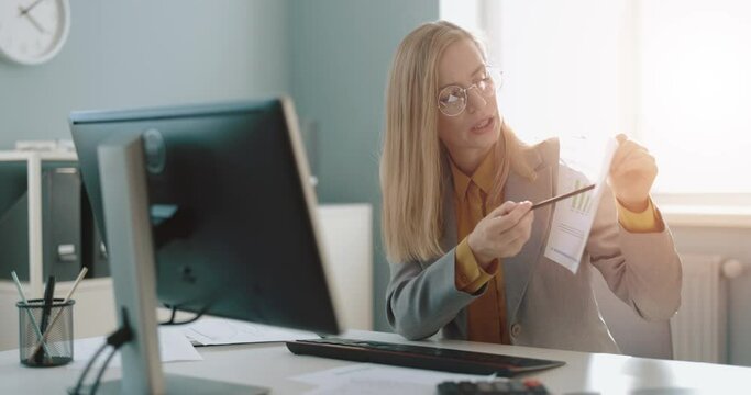 Caucasian woman in eyewear and formal clothes showing financial report during video chat at office. Confident lady sitting at desk and using moden pc for online meeting.