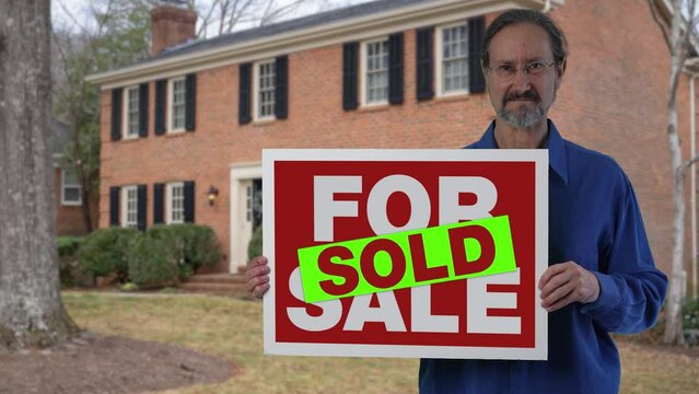 Happy smiling real estate agent holding For Sale SOLD sign in front of a house.