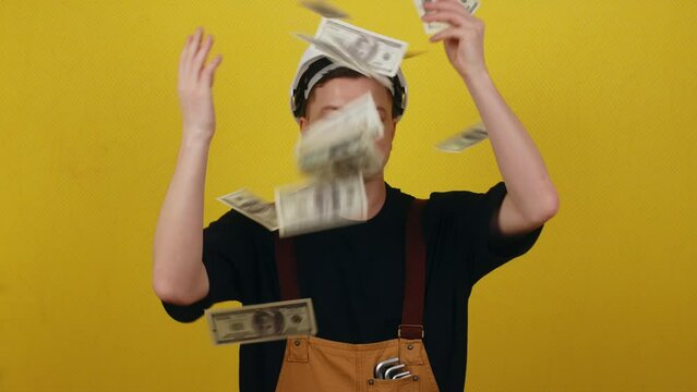 Portrait of a cheerful young builder in a hard hat throwing money on a yellow background