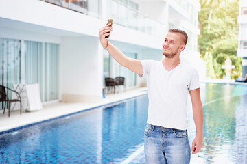 Fototapeta na wymiar Vacation and technology. Handsome young man using smartphone taking selfie in swimming pool.