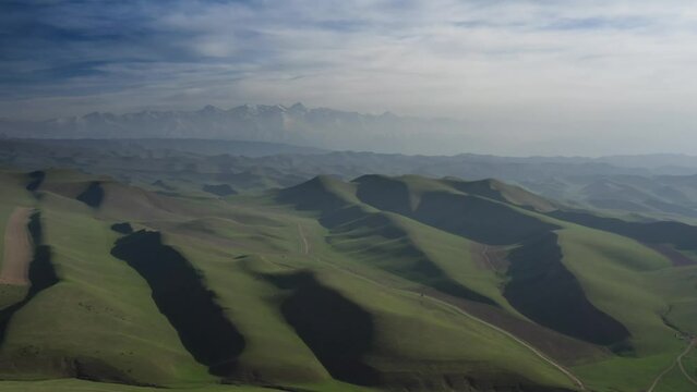 Amazing aerial footage of green hills with contrast light and blue mountains with ice peaks in the distance. Drone camera view of terrain in the highlands of warm green color overviewing snow caps.
