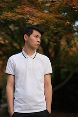 Fototapeta na wymiar one handsome Chinese young man in white polo shirt looking away with blur maple tree 