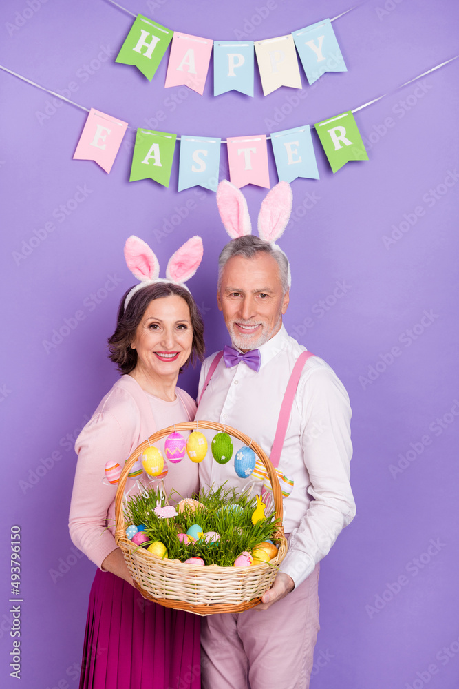 Sticker Vertical photo of two elderly cheerful people hold eggs basket bunny ears isolated on violet color background