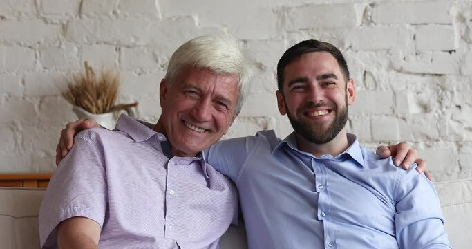 Head shot portrait multi generational handsome men, older father his young adult son hugging relaxing on couch feel overjoyed, spend weekend together, express care, looking happy. Family bond concept