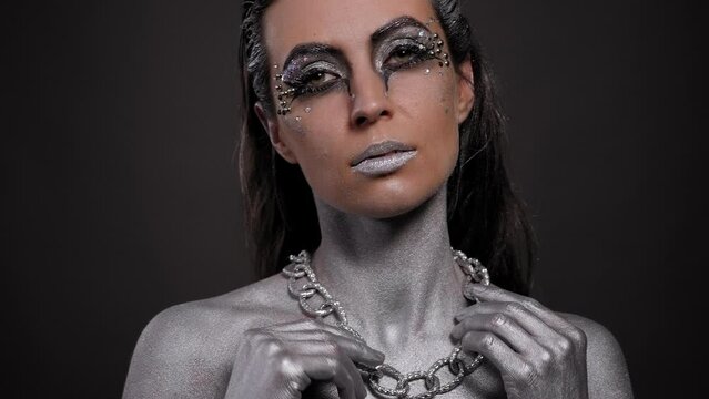 A gorgeous female model with silver body art on her body, bright makeup and silver hair poses for the camera, a chain hanging around her neck. Fashion.