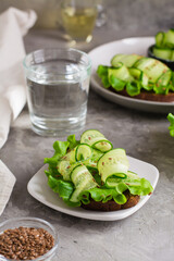 Appetizing bruschetta from rye bread, lettuce, cucumber and flax seeds on a plate and a glass of water on the table. Healthy diet food. Vertical view