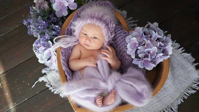 Newborn girl dressed in woolen cap and garment lies in heart-shaped wooden basket among purple flowers. Photo-shoot of cute baby in studio close upper view