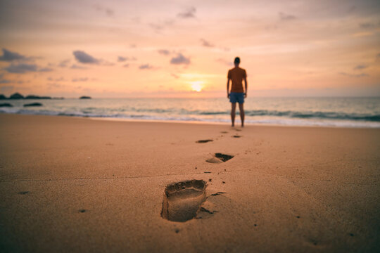 Footprints in sand against silhouette of person. Lonely man walking along beach to sea at golden sunset.