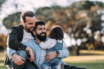 Male gay family embracing and happy in the park with their little daughter