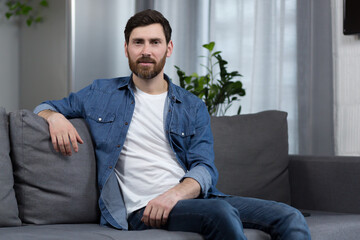 Portrait of a man with a beard at home, sitting on the couch and looking at the camera