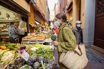 Woman in medical mask choosing fresh products at market stall on the famous gastronomical street in Bologna. Concept of buying local food during pandemic. Idea of Italian lifestyle - obrazy, fototapety, plakaty