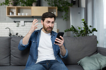 Frustrated and angry man looking at phone screen sitting on sofa at home