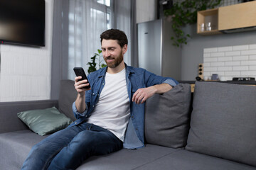 Fototapeta premium Happy man smiling and using phone sitting on sofa at home