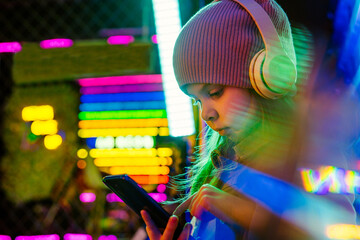 Little girl wearing headphones using smartphone on the colorful neon