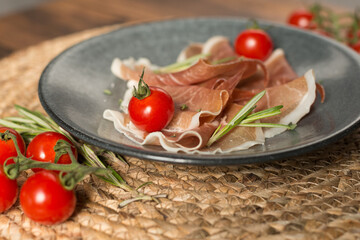 Dark plate with parma ham, ripe tomatoes, rosemary with salt. On wooden table.