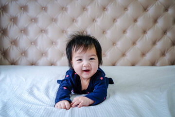 Cute little baby girl, playing at home in bed. Cute little caucasian baby lying on bed at home. Little cute baby girl, child in blue dress, smiling happily at camera in white sunny, bright bedroom.