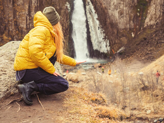 A woman in a yellow jacket squatting on the background of autumn nature put her hand to a waterfall flowing in the rocks in the distance. Soft focus. Optical illusion