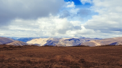 Fototapeta premium Brown autumn field in the highlands of the Caucasus overlooking a mountain valley and lush clouds in the blue sky