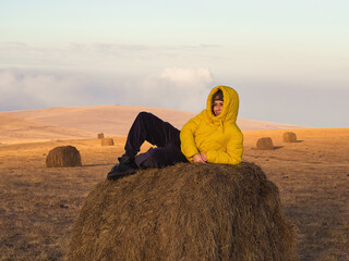 A smiling woman in a padded jacket lies on a haystack in an autumn field under the evening sky. Caucasian countryside