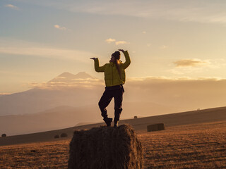 A woman stands in a pose on a haystack in a golden field against the backdrop of Elbrus seen from...