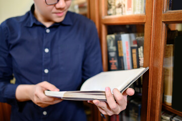 Smart Asian man university student reading book by vintage bookshelf. Textbook resources in college library for educational subject and research. Scholarship for education opportunity.