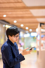 Smart Asian man wearing glasses using smartphone in shopping mall or department store. Telecommunication network and technology. Mobile phone app for urban lifestyle. 