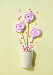 Pink lollipops in the shape of hearts at  paper cup with color sugar on a yellow background. Top view and copy space. Vertical orientation