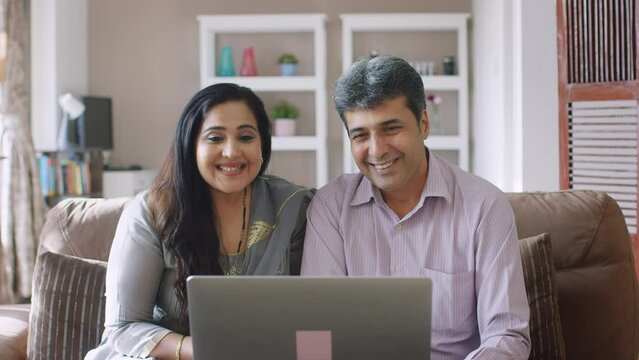 Happy Indian Asian Modern Middle-aged Retired Couple, Family Or Husband Wife Sitting Together In Front Of Laptop Waving Hands Interacting Over Online Video Call And Smiling In A Interior House Setup. 