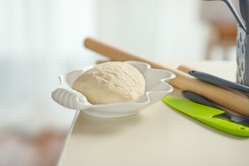 Kneaded dough in a white bowl on a kitchen table with kitchen utensils