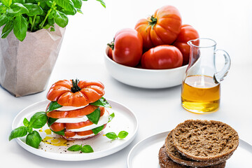 Tomato with mozzarella and basil. Caprice salad as a tower on white plate. Set table with bread and olive oil..