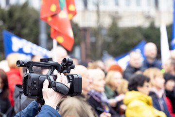 Filming street protest, television camera lens in the focus, blurred people in the background