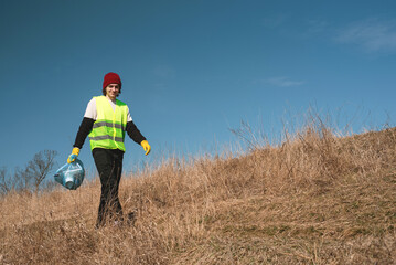 Naklejka na ściany i meble Man nature activist in yellow vest carries blue trash bag along the pathway in the sunny field