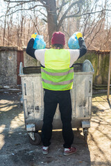 Man in yellow vest throws two blue trash bags out in the dumpster behind back photo