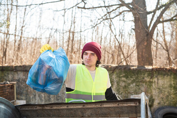 Man in yellow vest throws blue trash bag out in the dumpster