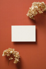 Blank white paper card mockup with dried flowers on marsala color background. Flat lay, top view, copy space.