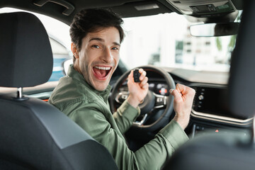 Excited Caucasian guy holding car key and gesturing YES, sitting in new auto at dealership store