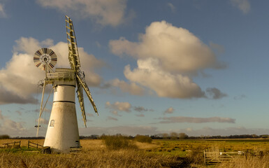 Iconic view of a restored historic Norfolk drainage mill, with sails on a sunny Spring morning. Set in flat fields, with blue sky and cloud formations. Landscape image with space for copy. Thurne, UK - 493831305
