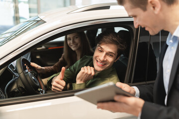 Young couple visiting car salon, sitting inside automobile, talking to sales assistant, looking at tablet in dealership