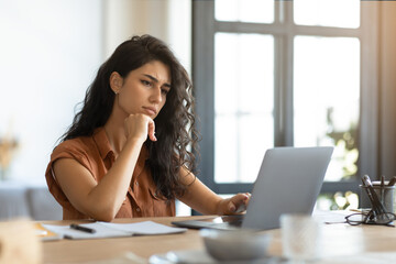 Thoughtful young brunette woman looking at laptop screen, working online at desk in home office,...
