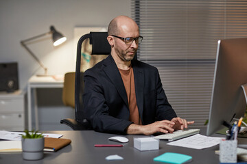 Portrait of bald adult businessman using computer at workplace while working late at night in office