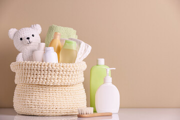 Knitted basket with baby cosmetic products, bath accessories and toy bear on white table against beige background. Space for text