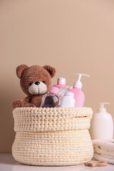 Knitted basket with baby cosmetic products and toy bear on white table against beige background
