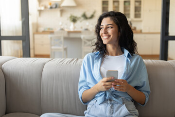 Portrait of smiling young woman with smartphone sitting on couch at home, using mobile device, copy space