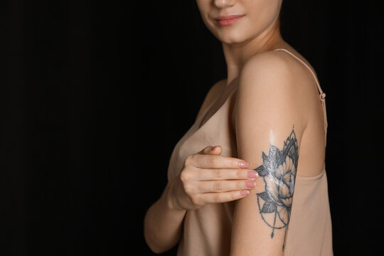 Woman applying cream onto her arm with tattoo on dark background, closeup. Space for text