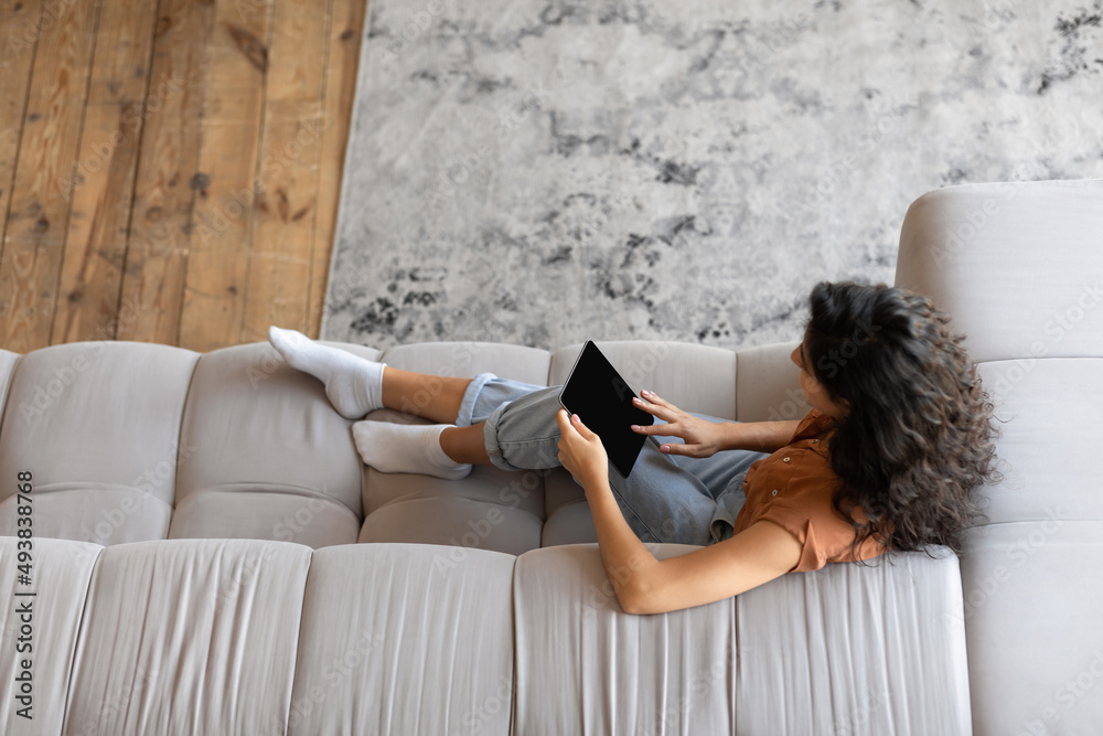 Poster top view of young woman with tablet computer sitting on sofa, working or studying online from home, 
