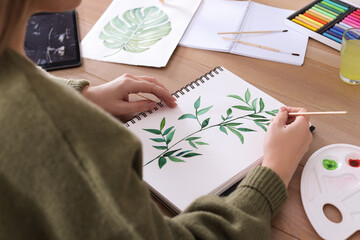 Woman painting green twig in sketchbook at wooden table, closeup