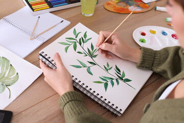 Woman painting green twig in sketchbook at wooden table, closeup